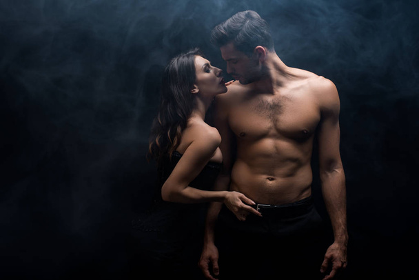 Seductive woman touching belt of muscular man on black background with smoke - Photo, image