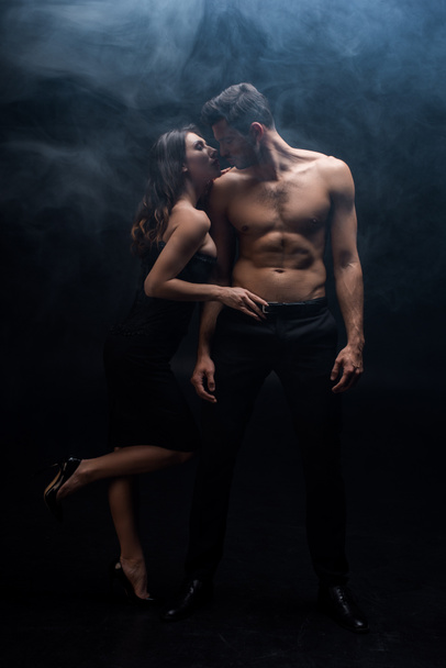 Full length of sensual woman touching belt of muscular man on black background with smoke - Photo, Image