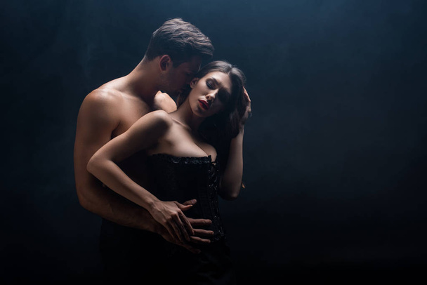 Πλευρική άποψη του όμορφου γυμνόστηθη άντρας αγκαλιάζει σέξι γυναίκα απομονωμένη σε μαύρο - Φωτογραφία, εικόνα