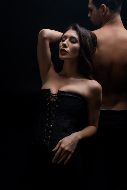 Sexy vrouw in korset staan door shirtless man geïsoleerd op zwart - Foto, afbeelding