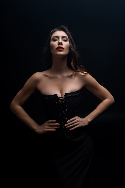 Sexy woman in corset with hand on hips looking at camera isolated on black - Фото, изображение