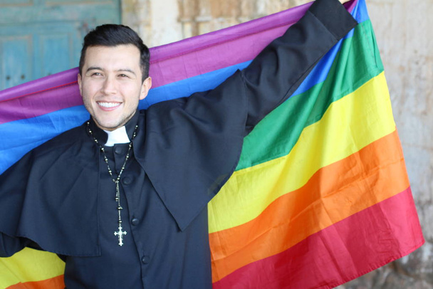 Smiling handsome priest in cassock holding rainbow LGBT flag posing on shabby church background - Foto, imagen