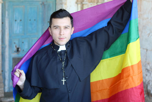 Serious priest in cassock holding rainbow LGBT flag posing on church background - Фото, изображение