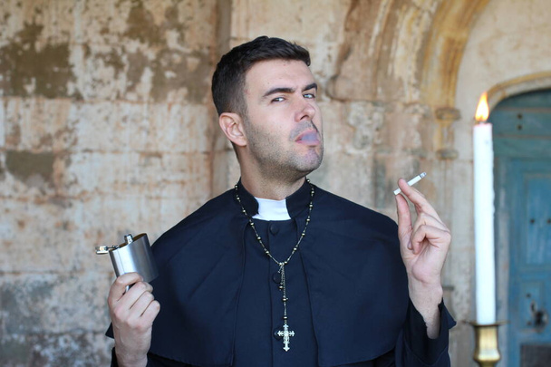 Priester raucht Zigarette und trinkt Alkohol, posiert vor dem Hintergrund der Kirche mit brennender Kerze - Foto, Bild