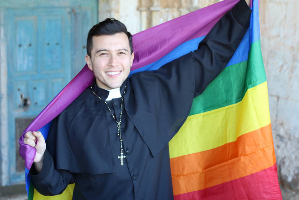 Улыбающийся красивый священник в рясе с радужным ЛГБТ-флагом, позирующий на потрепанном церковном фоне
 - Фото, изображение