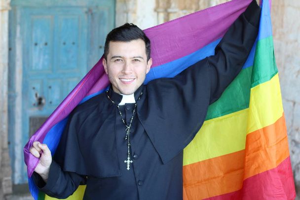 Smiling handsome priest in cassock holding rainbow LGBT flag posing on shabby church background - Foto, afbeelding