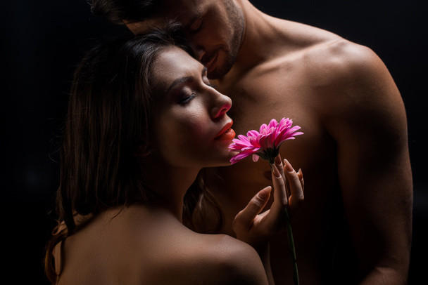Beautiful woman holding gerbera by handsome shirtless man isolated on black  - Fotó, kép