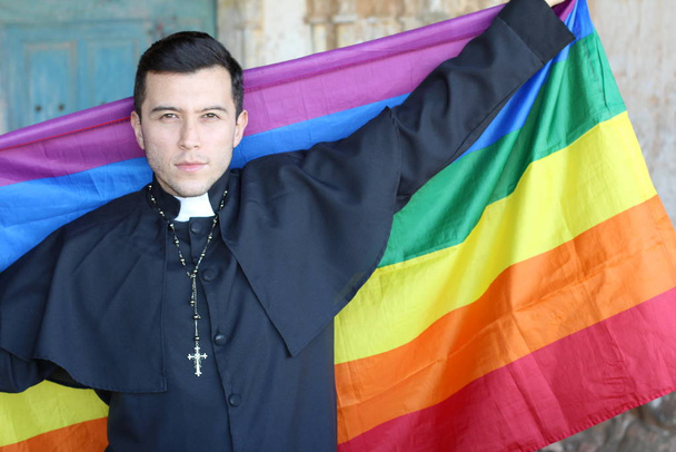 Serious priest in cassock holding rainbow LGBT flag posing on church background - Foto, immagini