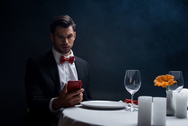 Elegante uomo che guarda smartphone al tavolo servito con candele su sfondo nero con fumo
  - Foto, immagini