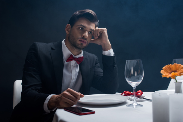 Élégant homme avec la main par la tête assis par smartphone sur une table servie sur fond noir avec de la fumée
  - Photo, image