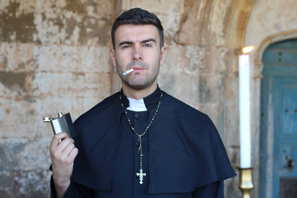Sacerdote fumar cigarrillo y beber alcohol posando en el fondo de la iglesia con vela encendida
 - Foto, imagen