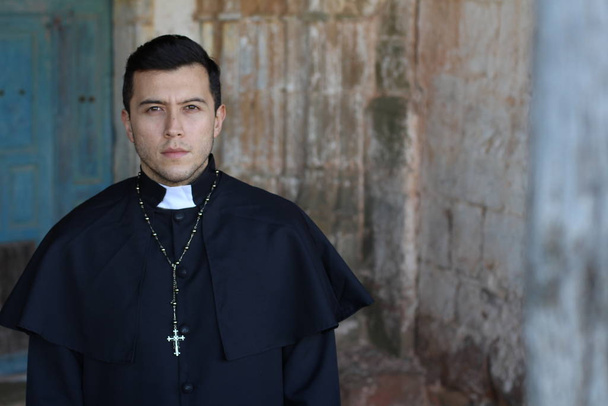 Vakava pappi kasukassa poseeraa nuhjuisella kirkon taustalla
 - Valokuva, kuva