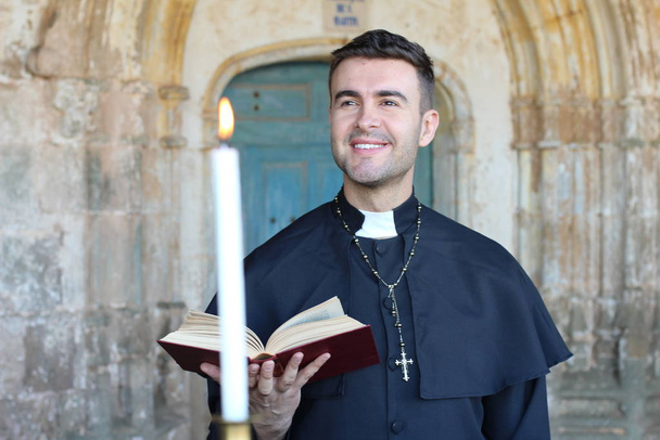 Улыбающийся священник с открытой библией на церковном фоне
 - Фото, изображение