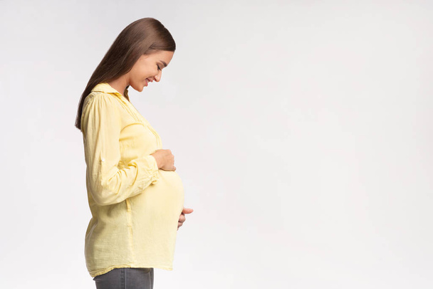 Νεαρή έγκυος γυναίκα που αγγίζει την κοιλιά στέκεται σε λευκό φόντο, πλευρική άποψη - Φωτογραφία, εικόνα