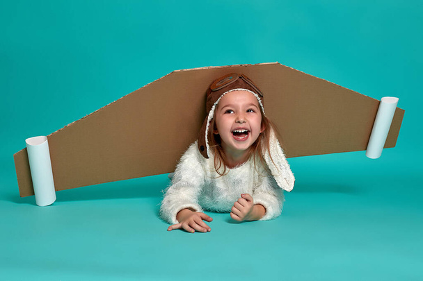Dziewczynka bawi się skrzydełkami na niebieskim tle. Szczęśliwe dziecko w kostiumie astronauty, marzące o zostaniu astronautą. Portret dziecka. - Zdjęcie, obraz