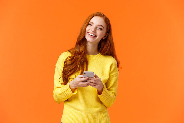 Girl searching gifts for holidays online. Carefree happy redhead female student using smartphone app, laughing joyfully, holding mobile phone, chatting or browsing social networks, orange background - Photo, Image