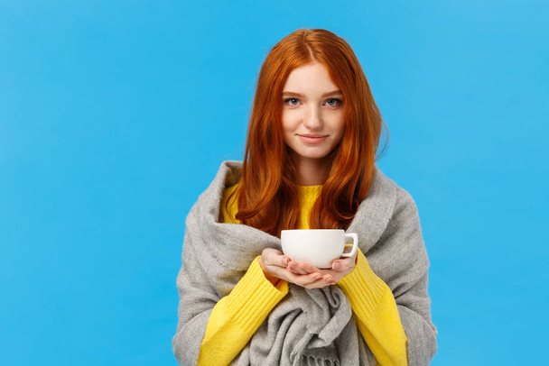 Redhead girl trying get warm, feeling cold wrapping herself with scarf on shoulders, drinking hot tea, holding teapot and smiling lovely, standing tender and cute blue background - Fotografie, Obrázek