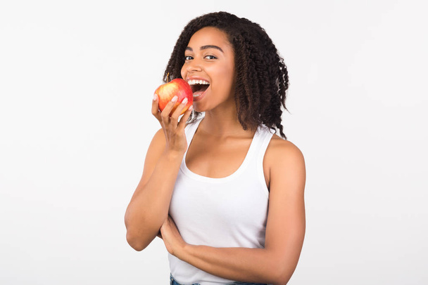 Χαρούμενο μαύρο κορίτσι που τρώει μήλο κοιτάζοντας την κάμερα - Φωτογραφία, εικόνα