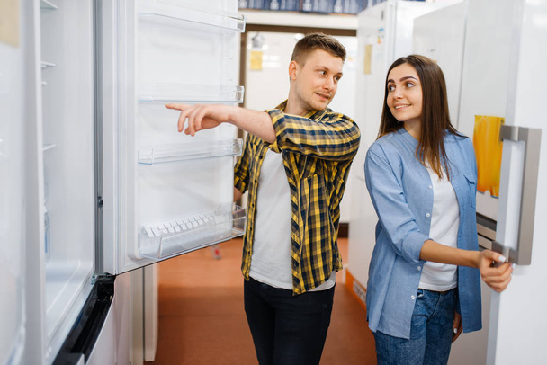 Casal jovem família escolhendo geladeira na loja de eletrônicos. Homem e mulher comprando eletrodomésticos no mercado
 - Foto, Imagem