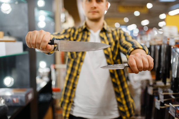 Ο νεαρός κρατάει μαχαίρια κουζίνας στο κατάστημα ειδών οικιακής χρήσης. Άνδρας που αγοράζει είδη οικιακής χρήσης στην αγορά, τύπος στο κατάστημα ειδών κουζίνας - Φωτογραφία, εικόνα