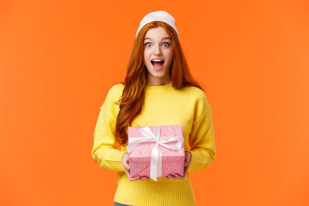 Surprised and excited happy girl unwrapping gifts on christmas eve, new year celebration, smiling amused and thrilled, holding wrapped box present, wearing winter beanie, orange background - Photo, Image