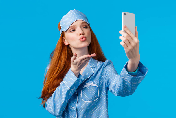 Portret w pasie czarującej rudej kobiety w ładnej piżamie, mówiącej "dobranoc", uzależnionej od telefonu, robiącej selfie, całującej się w powietrze przed kamerą, stojącej na niebieskim tle " - Zdjęcie, obraz