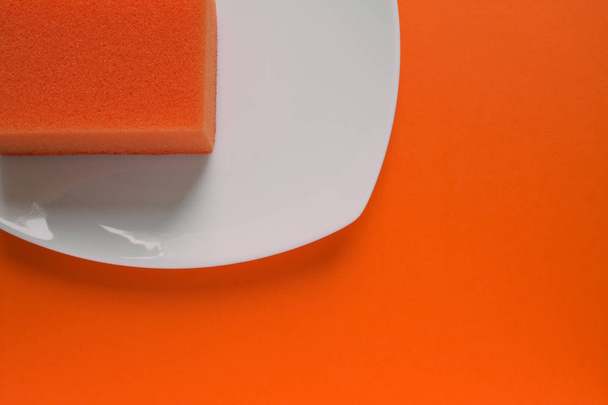 Orange sponge for washing dishes on a square white plate close-up on a plain background. Concept: clean dishes - Zdjęcie, obraz
