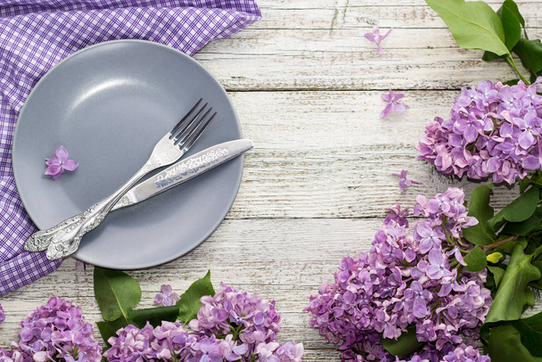 spring table setting with plate fork and knife decorated with lilac flowers on white wooden background - Photo, Image