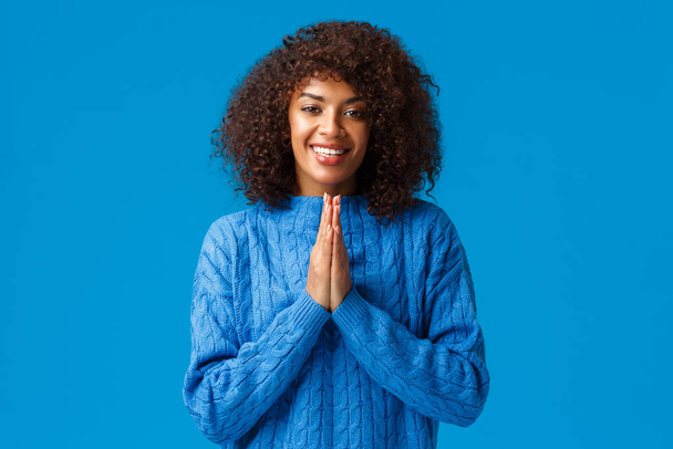 Dankbar süß lächelnde afrikanisch-amerikanische hübsche Frau mit Afro-Haarschnitt, sagt arigato und lächelt, klammert die Hände vor der Brust zusammen und sieht dankbar aus, zeigt Respekt, steht auf blauem Hintergrund - Foto, Bild
