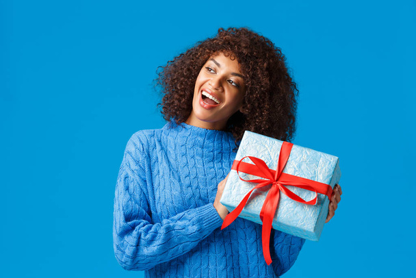好奇心と興奮した幸せな若いアフリカ系アメリカ人の女性は、クリスマス休暇のための贈り物を受け取り、青い背景に立って興味をそそる立って、内側に何を推測するために現在握手 - 写真・画像