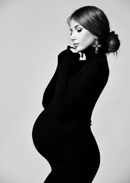 Young beautiful pregnant woman in black dress and massive earrings standing and looking down  - Фото, зображення