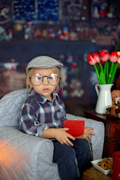 Sweet toddler boy, wearing glasses, reading a book and drinking  - Fotó, kép