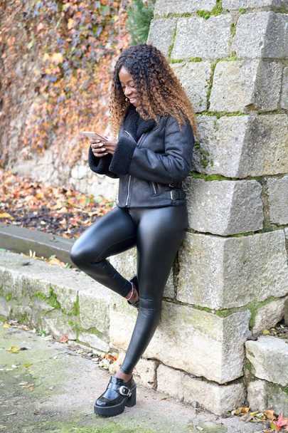 glückliches schwarzes Afro-Mädchen in Schwarz mit lockigem braunem Haar lächelt, während es mit seinem Handy auf Online-Informationen zugreift - Foto, Bild