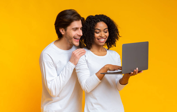 Межрасовые супруги держат ноутбук и с волнением смотрят на экран компьютера
 - Фото, изображение