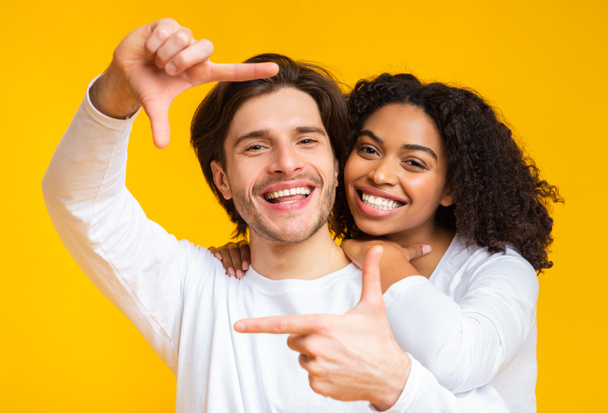 Positive multiracial couple looking through finger frame, posing over yellow background - Photo, Image