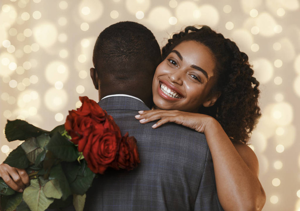 Χαρούμενη μαύρη γυναίκα που κρατάει κόκκινα τριαντάφυλλα ενώ αγκαλιάζει τον άντρα της - Φωτογραφία, εικόνα