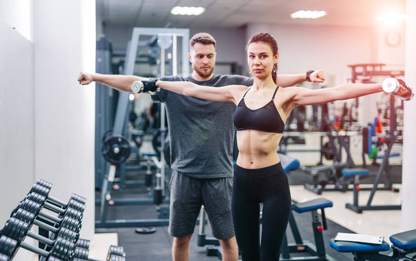 Smiling female in black top and leggings holding dumbbells before coach at gym. Male trainer and beautiful woman lifting metal dumbbells. - Photo, Image
