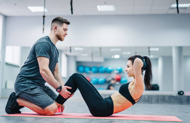 Young woman doing abdominal exercises with the help of her coach on a mat in gym. Muscular trainer helps attractive female doing abs - Photo, image