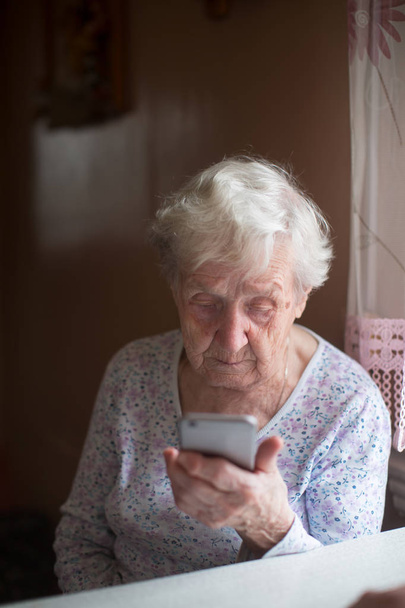 Stará důchodkyně sedí doma s chytrým telefonem v ruce. - Fotografie, Obrázek