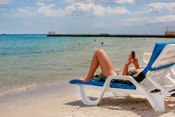 Dívka s chytrým telefonem na plastovém lehátku stojící na písku pláže na pozadí moře. - Fotografie, Obrázek