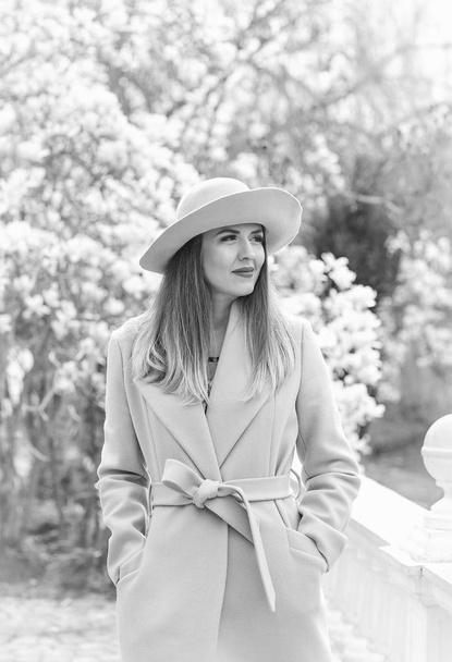 black and white photo of a young, beautiful woman, in a fashionable coat and hat, walking in the Park in the spring near a flowering Magnolia tree, on a Sunny day - Foto, imagen