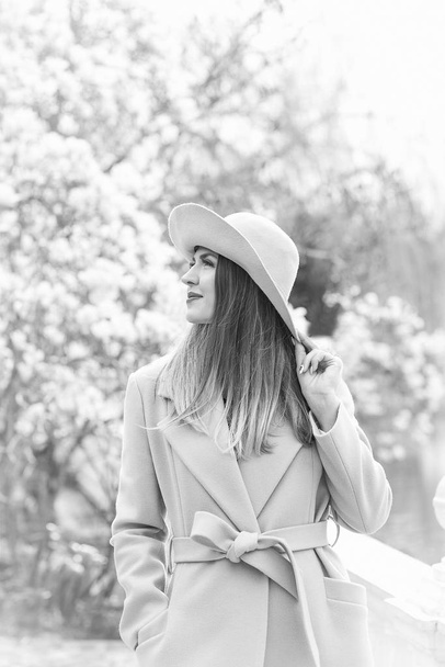 black and white photo of a young, beautiful woman, in a fashionable coat and hat, walking in the Park in the spring near a flowering Magnolia tree, on a Sunny day - Zdjęcie, obraz