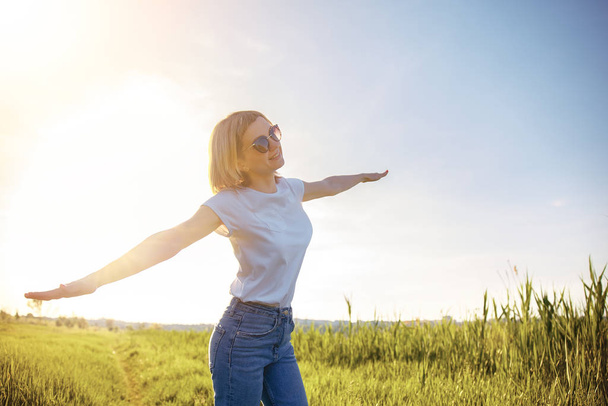 Beyaz tişörtlü, güneş gözlüklü ve kot pantolonlu kadın gün batımında uçağın taklidini yaparak kollarını ayırıyor. Yaz. Tatil hava yolculuğu konsepti - Fotoğraf, Görsel
