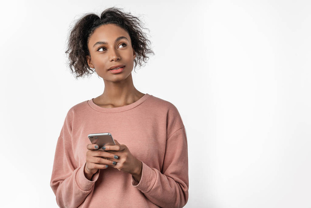 Nuoren afrikkalaisen naisen muotokuva, jolla on matkapuhelin seisoessaan ja katsoessaan pois valkoiselta taustalta
 - Valokuva, kuva