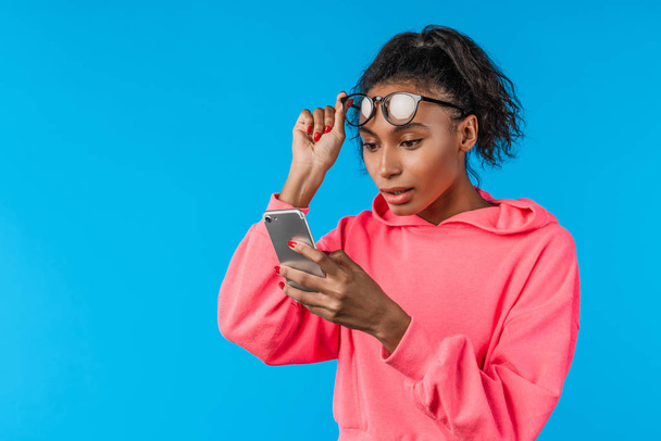 Meglepett fiatal afrikai nő szemüvegben tartja mobiltelefon felett kék háttér - Fotó, kép