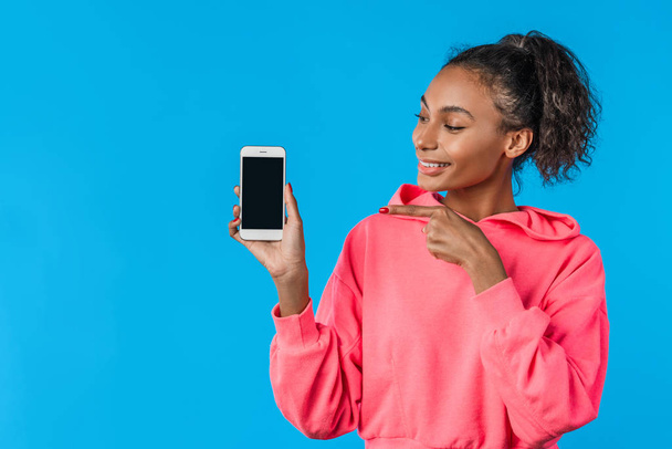 Cheerful young afro woman holding mobile phone and pointing finger over blue background - Foto, immagini