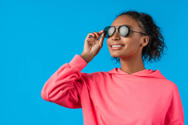 Beautiful african american female model wear sunglasses and posing over blue background - Fotoğraf, Görsel