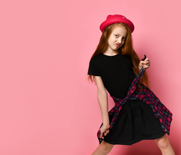 Teenage kid in black dress and red hat. She tightens checkered shirt on her waist while posing against pink background. Close up - Photo, Image