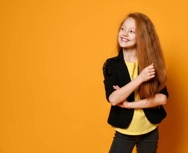 Ginger teen female in black jacket and pants, yellow t-shirt. She is smiling, hands folded, posing on orange background. Close up - Photo, image