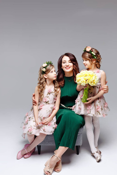 Beautiful happy family mom and two little cute girls in wreaths of fresh flowers and a large bouquet of tulips kiss, laugh and hug in the studio on a light background - Photo, image